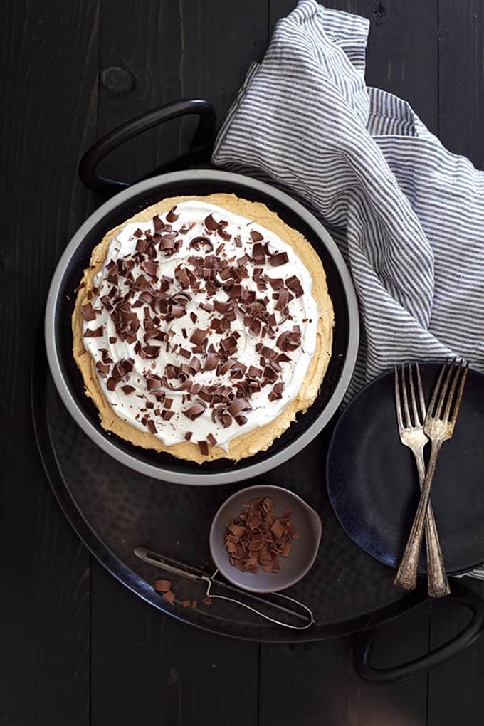 An overhead shot of a Black Bottom No Bake Pumpkin Cheesecake on a metal tray with black plates, two forks, a striped napkin and a small bowl of chocolate shavings with a vegetable peeler.