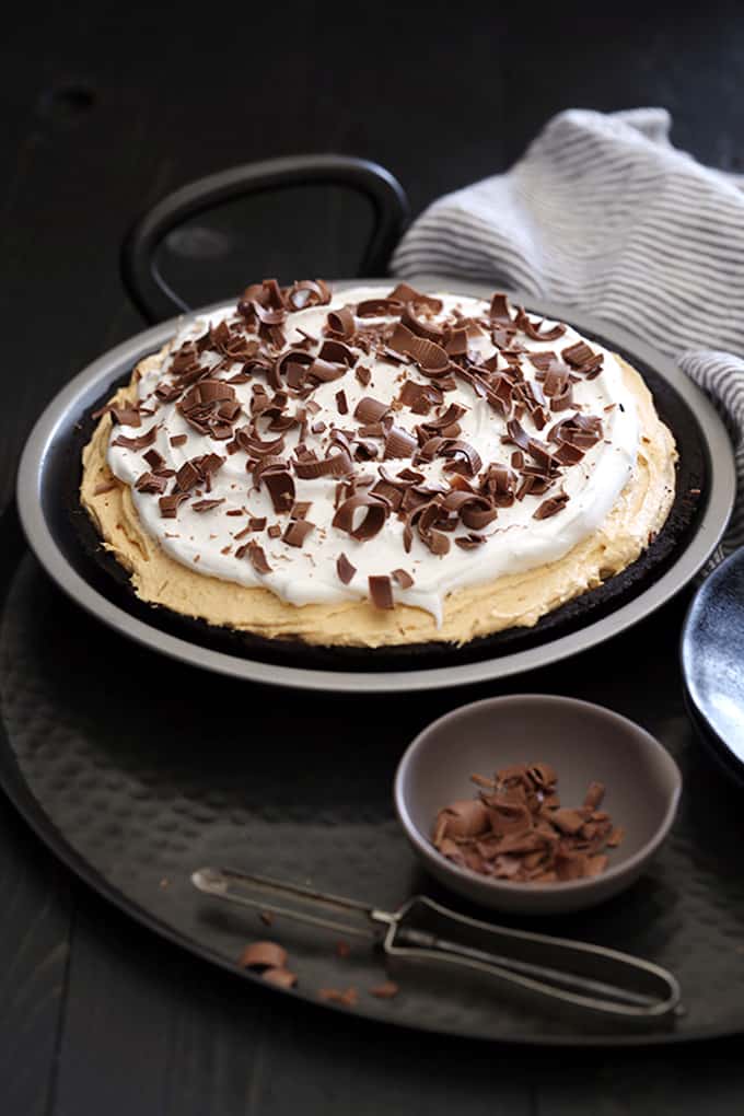 A Black Bottom No Bake Pumpkin Cheesecake sits on a metal tray with striped napkin and a small bowl of chocolate shavings with a vegetable peeler.