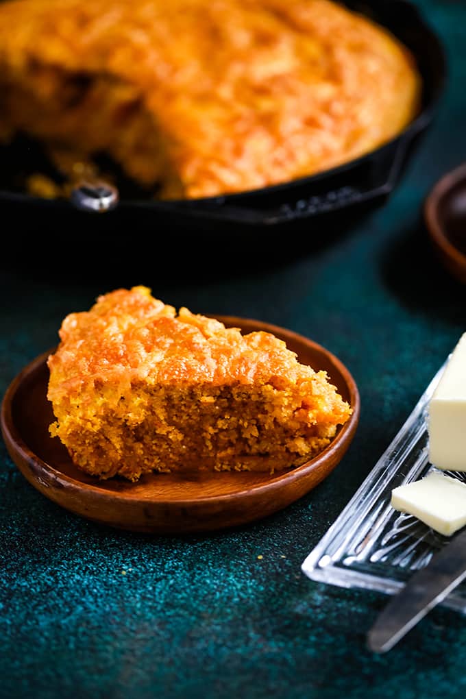 A wedge of Pumpkin Cream Cheese Skillet Cornbreads sits on a wooden plate on a green speckled background with a skillet of cornbread behind it.