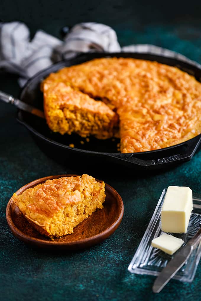 A skillet of Pumpkin Cream Cheese Skillet Cornbread with two wedges cut and the skillet sitting on a speckled green background.