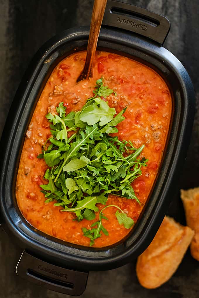 A slow cooker full of Slow Cooker Sausage Lentil and Arugula Soup sits on a dark background by two pieces of crusty bread.