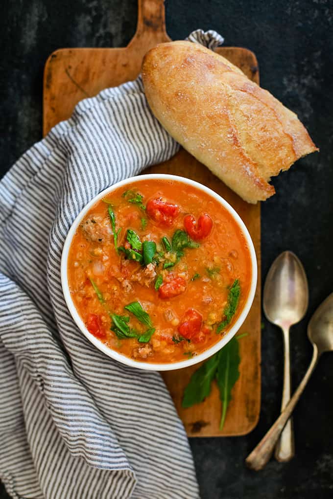 A white bowl of Slow Cooker Sausage Lentil and Arugula Soup sits on a wood cutting board next to a striped napkin, crusty bread and two spoons.