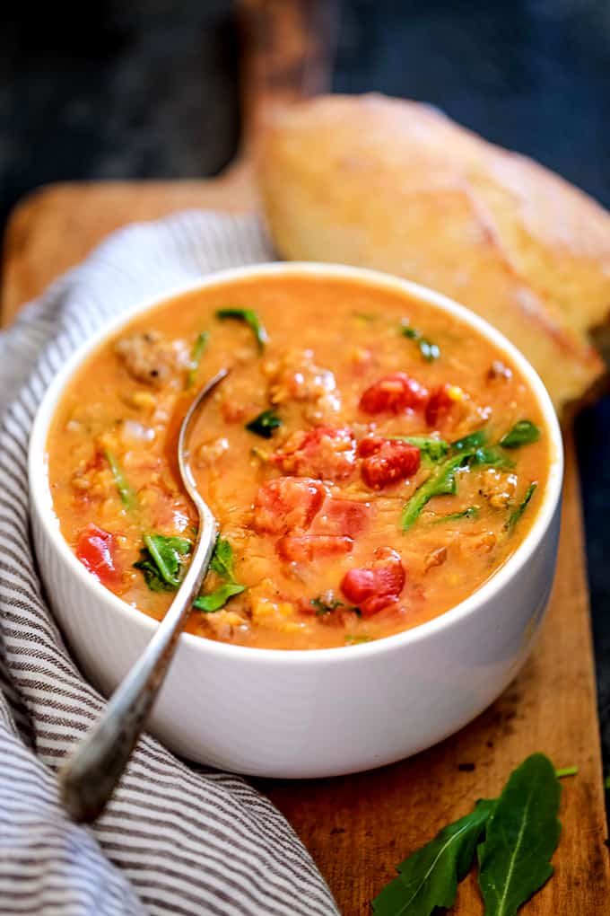 A white bowl of Slow Cooker Sausage Lentil and Arugula Soup sits on a wood cutting board next to a striped napkin, crusty bread and two spoons.