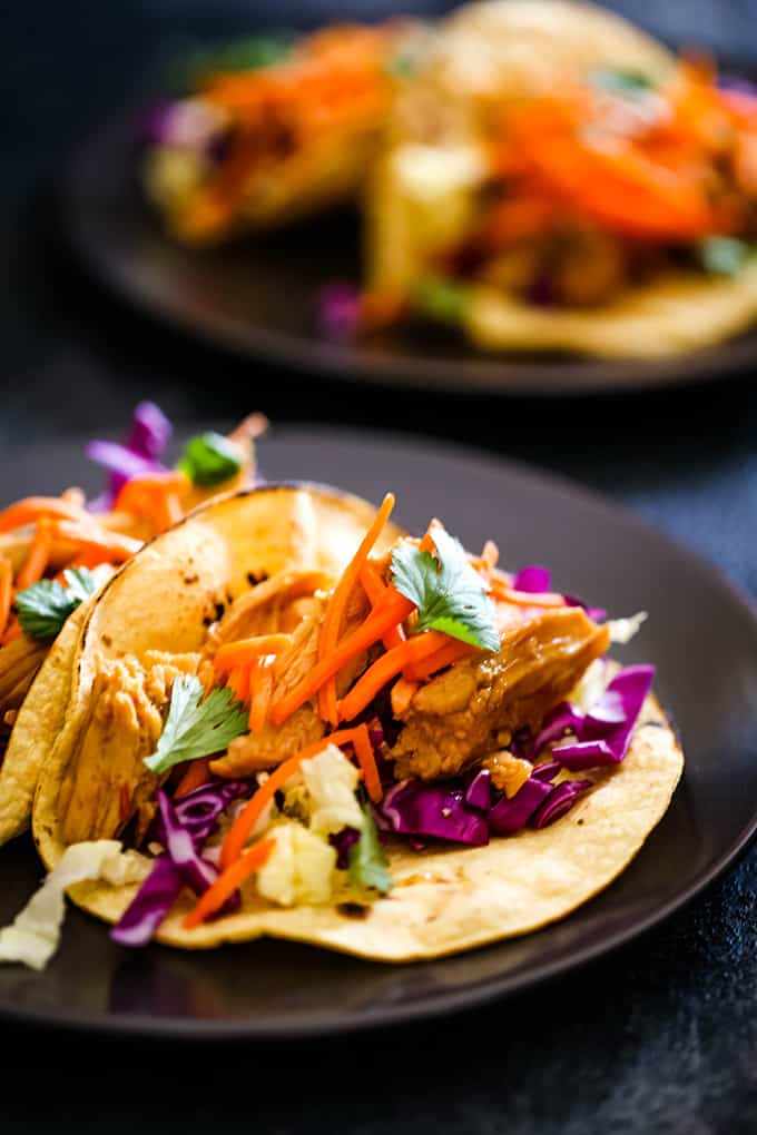 Slow Cooker Hoisin Chili Chicken Tacos are garnished with slaw and cilantro on dark colored plates.