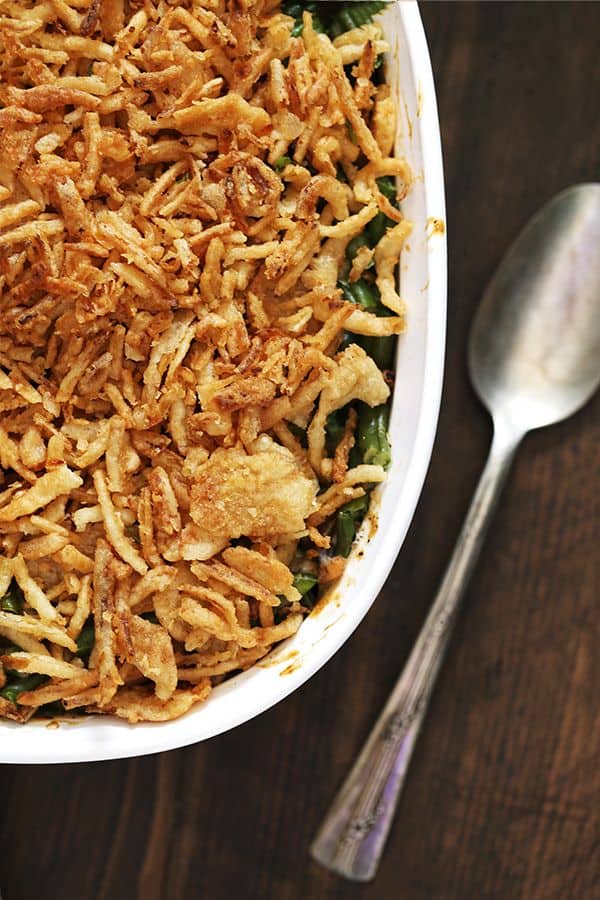 A white casserole dish of Twice the Onion Green Bean Casserole sits on a wooden surface with a spoon sitting to the right of it.