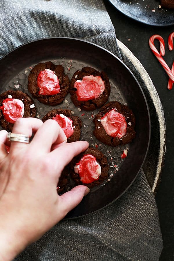 A hand reaches for a cookie on a platter of Mocha Peppermint Crunch Cookies sits on a grey napkin on a dark surface surrounded by a black plate with additional cookies, candy canes and more peppermint candy.