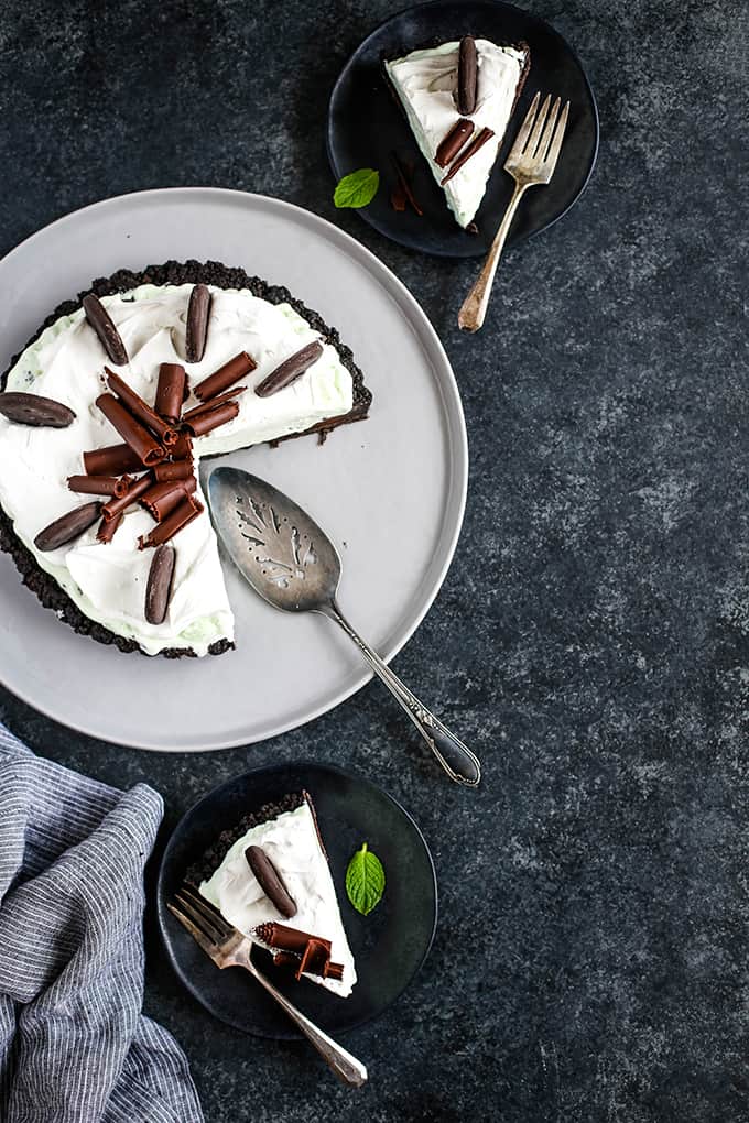 A Grasshopper Ice Cream Tart sits on a grey serving plate while two slices of tart rest on black plates with forks.