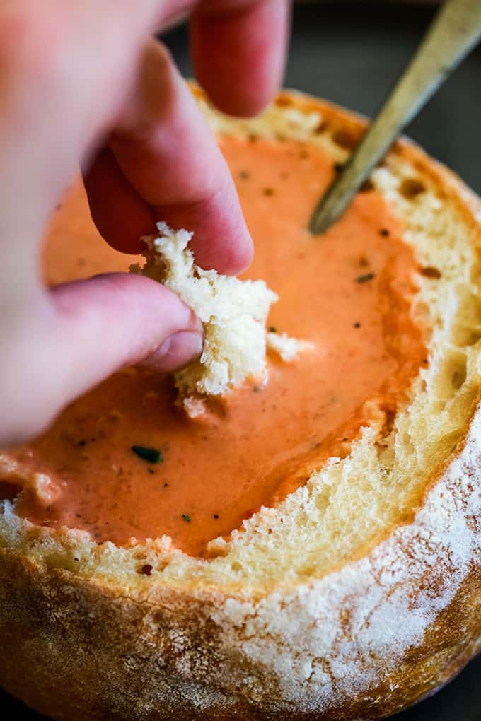 A hand dips a piece of bread into a bread bowl filled with Creamy Tomato Bacon Soup.