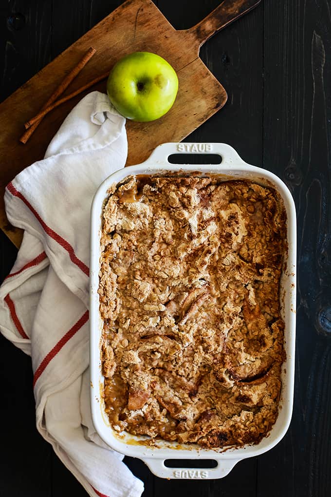 A white baking dish of Snickerdoodle Apple Crisp flanked by a white towel and wooden cutting board.