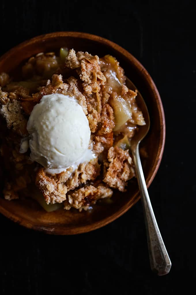 A serving of Snickerdoodle Apple Crisp on a wooden plate with a spoon and topped with a scoop of vanilla ice cream.