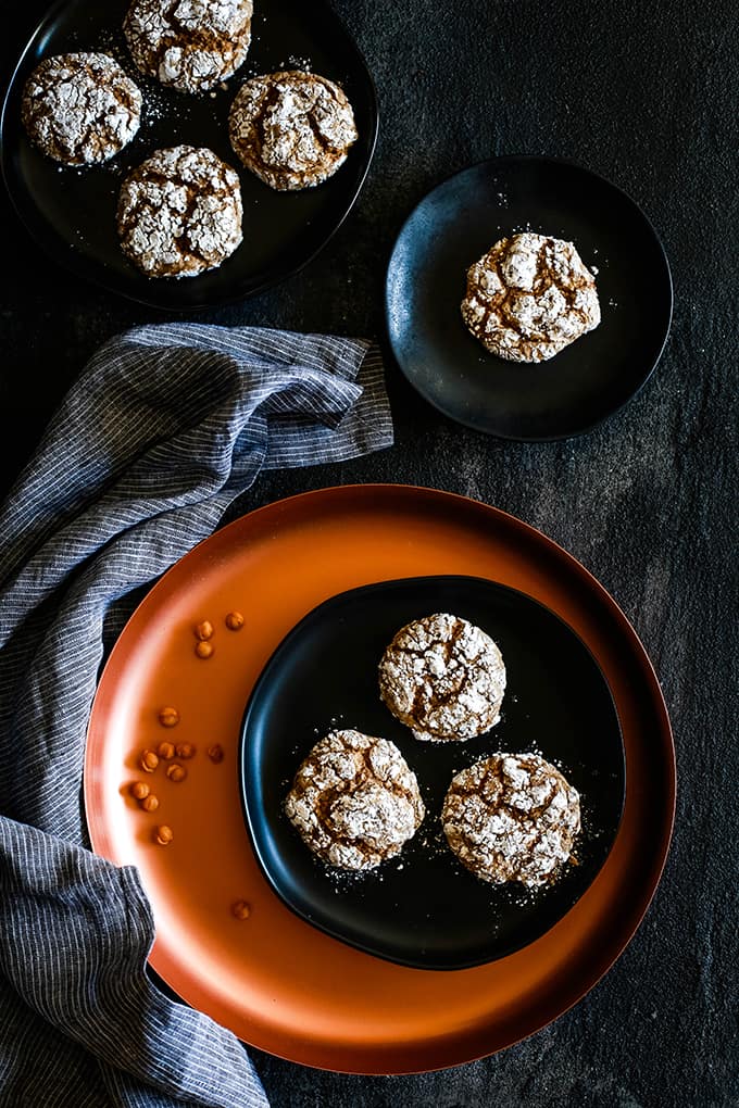 A stack of Caramel Pumpkin Spice Crinkle Cookies on a black background. #caramel #pumpkinspice #cookies #cookierecipes #melaniemakes Caramel Pumpkin Spice Crinkle Cookies on black plates on a black background. One plate is on a copper charger with caramel bits and a striped napkin sits to the left.