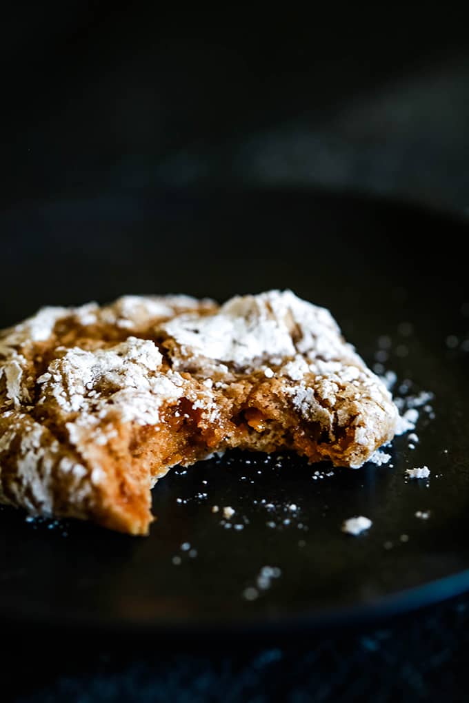 A stack of Caramel Pumpkin Spice Crinkle Cookies on a black background. #caramel #pumpkinspice #cookies #cookierecipes #melaniemakes A Caramel Pumpkin Spice Crinkle Cookie with a bite removed sitting on a black plate.