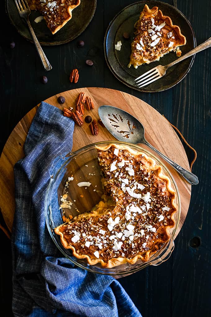 An overhead shot of a Chocolate Pecan Coconut Custard Pie on a wooden platter surrounded by a blue napkin, pie server, pecans, chocolate chips and two plates with pieces of pie and forks.