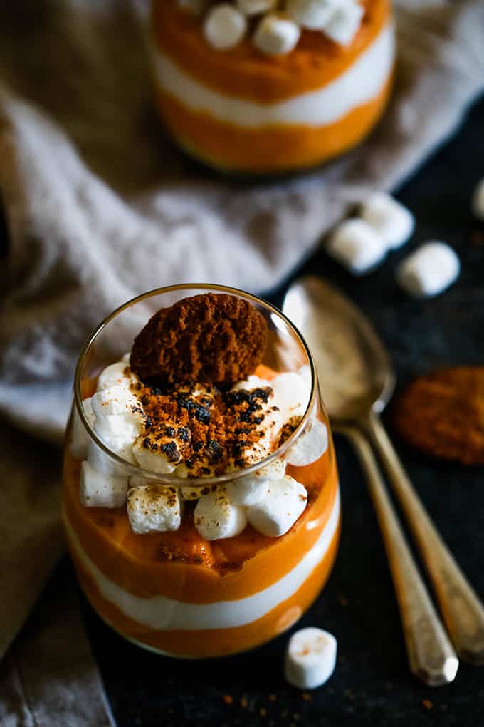 A glass of Gingersnap Sweet Potato Mousse sits on a dark surface surrounded by two spoons, mini marshmallows and a neutral colored background. An additional dessert sits in the background.