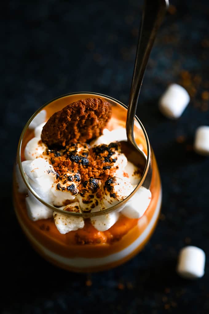 A glass of Gingersnap Sweet Potato Mousse with a spoon in the top sits on a dark surface surrounded by three mini marshmallows.