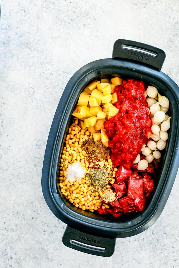 A slow cooker insert is filled with the necessary ingredients to create Slow Cooker Mexican Beef Stew and sits on a white speckled background.