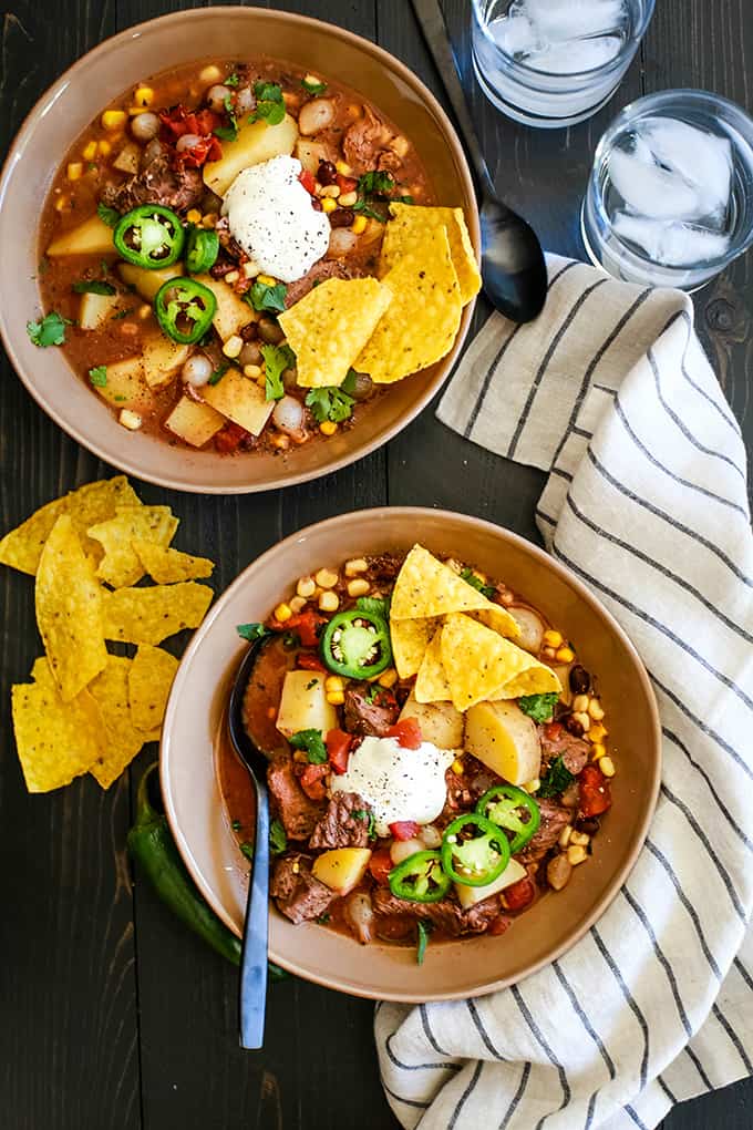 Two bowls of Slow Cooker Mexican Beef Stew sit on a dark background surrounded by a striped napkin, two glasses of water and yellow tortilla chips.