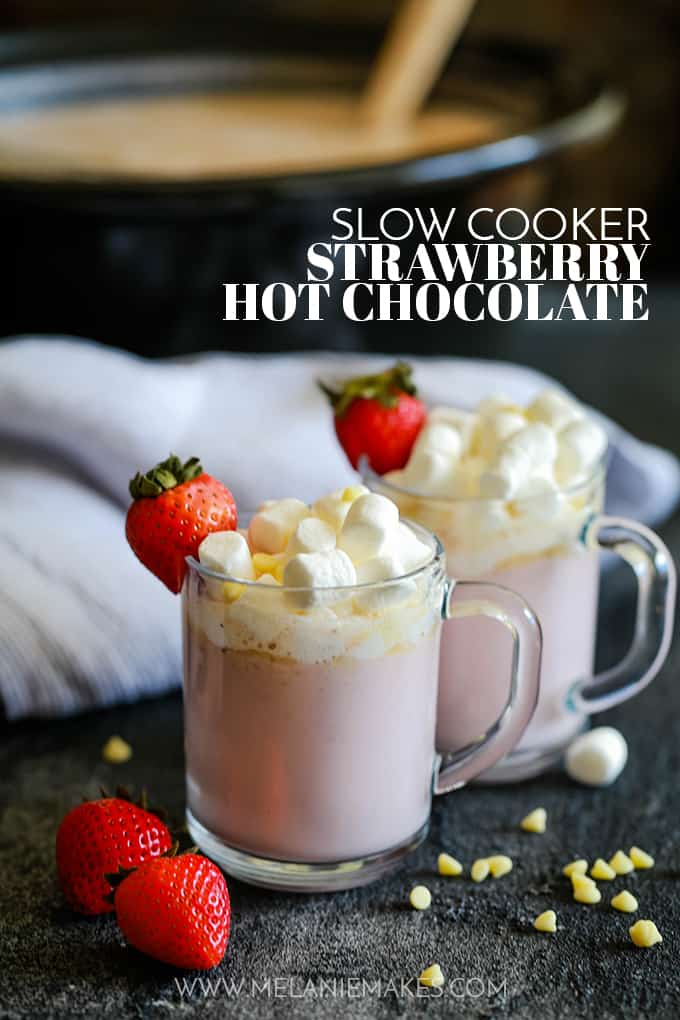 Two mugs of Slow Cooker Strawberry Hot Chocolate are topped with marshmallows, white chocolate and a strawberry and sit on a black surface. Behind them are a striped napkin and slow cooker full of hot chocolate.
