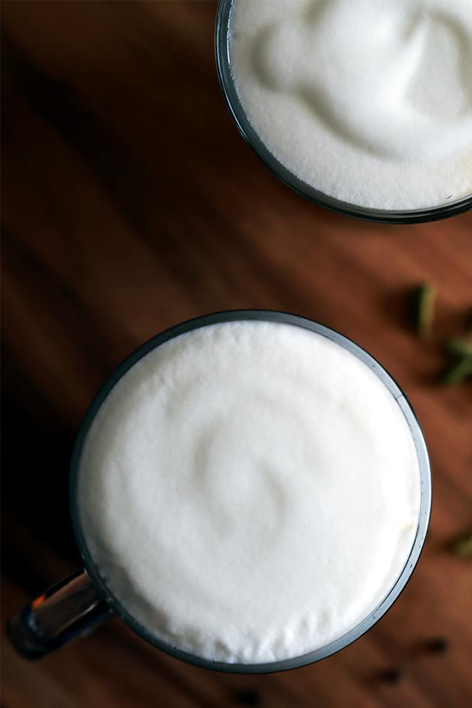 Two mugs of chai latte made from Homemade Chai Tea Concentrate sit on a wooden tray on a dark background surrounded by spices.