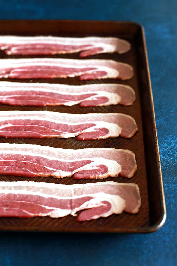 This Oven Roasted Bacon is the easiest and less messy way to make bacon. EVER. Forget spattering grease and standing over a screaming hot stovetop. In just 15 minutes, you can have perfectly crisp bacon with hardly any effort! #bacon #breakfast #roasted #oven #pork #brunch Six slices of uncooked bacon are arranged in horizontal rows on a baking sheet sitting on a dark blue background.