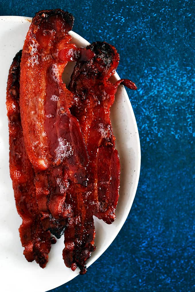 This Oven Roasted Bacon is the easiest and less messy way to make bacon. EVER. Forget spattering grease and standing over a screaming hot stovetop. In just 15 minutes, you can have perfectly crisp bacon with hardly any effort! #bacon #breakfast #roasted #oven #pork #brunch Four slices of Oven Roasted Bacon sit on a white plate on a dark blue background.