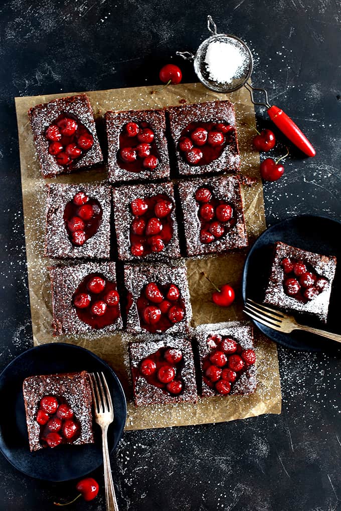 Fudgy Black Forest Bars sit on a brown parchment square on a black background. Two bars are on black plates with forks and powdered sugar rests in a sifter.
