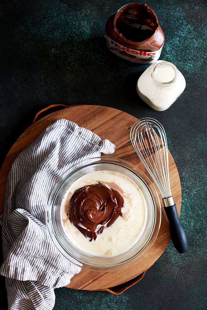 A glass bowl of Microwave Nutella Ganache sits on a wooden tray with a whisk and napkin and a jar of Nutella and a bottle of cream.