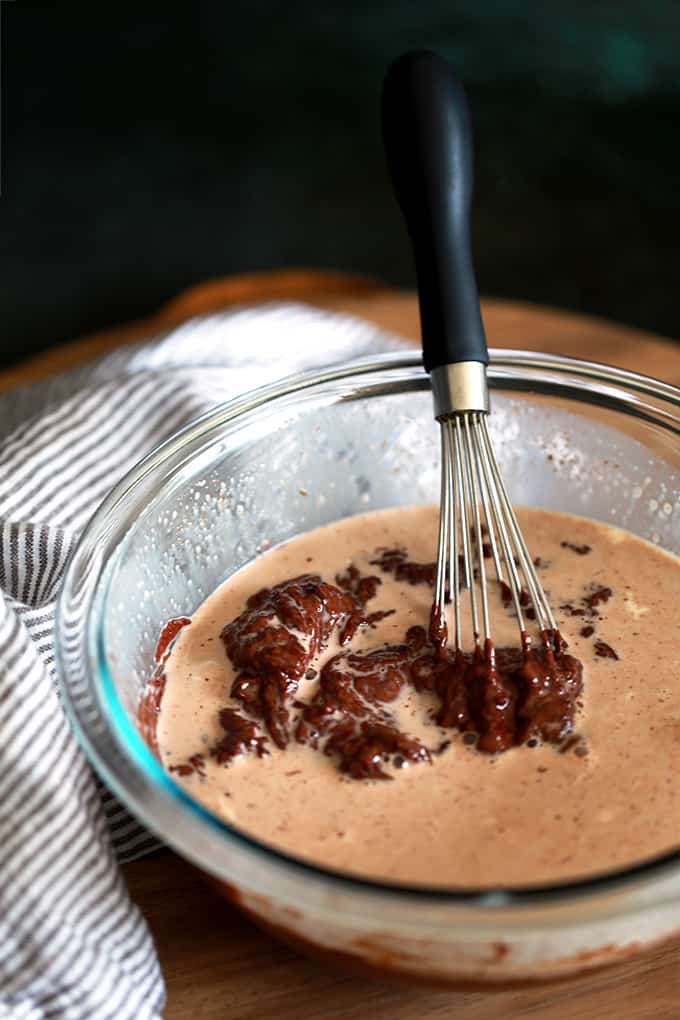 A glass bowl of Microwave Nutella Ganache with a whisk sits on a wooden tray with a striped napkin.