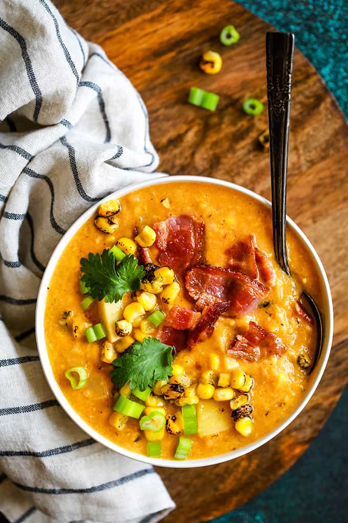 Slow Cooker Chipotle Pumpkin Corn Chowder garnished with bacon, corn, cilantro and green onions in a white bowl on a wooden plate with a spoon in the chowder.