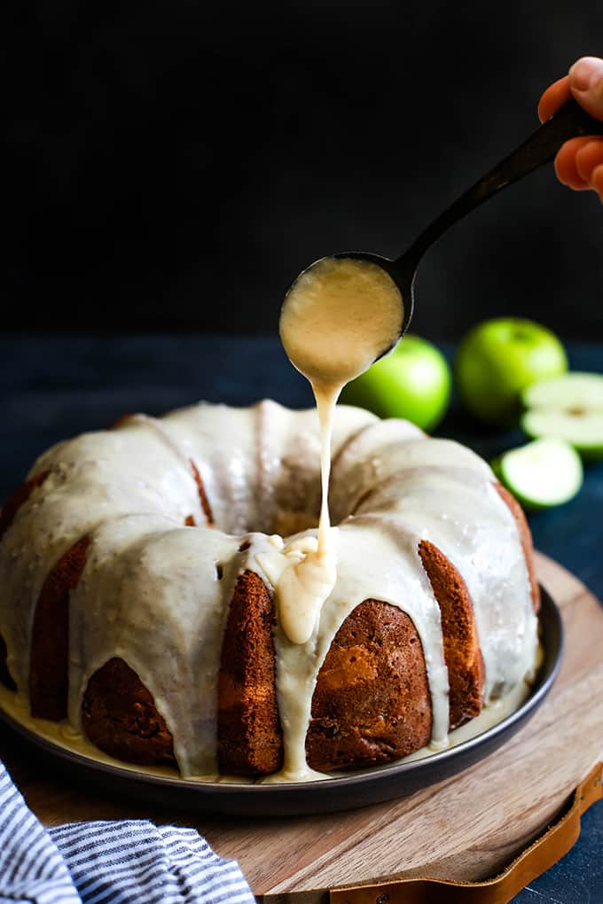 An Apple Cream Cheese Bundt Cake is being drizzled with caramel icing on a plate sits atop a wood serving board flanked by a striped towel and green apples.