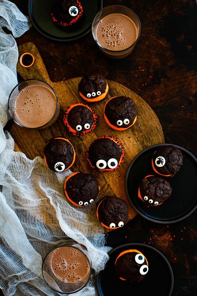 An overhead view of Mini Monster Chocolate Whoopie Pies on a wooden board and black plates with glasses of chocolate milk.