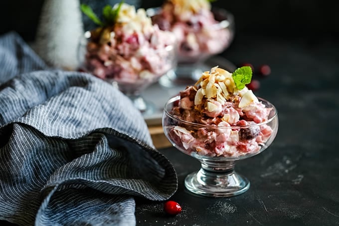 A footed dish of Cranberry Millionaire Salad sits on a dark background with a striped napkin alongside and additional dishes in the background.