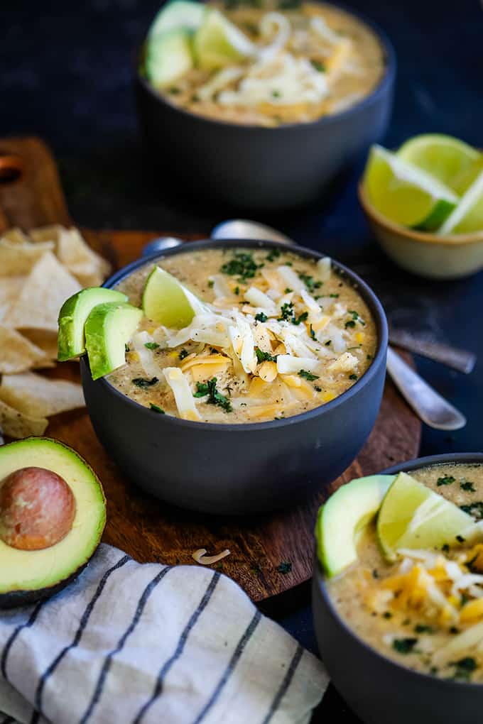 Three bowls of Slow Cooker Coconut Lime White Chicken Chili sit on a wooden surface surrounded by tortilla chips, an avocado half, a striped napkin and a bowl lime wedges.