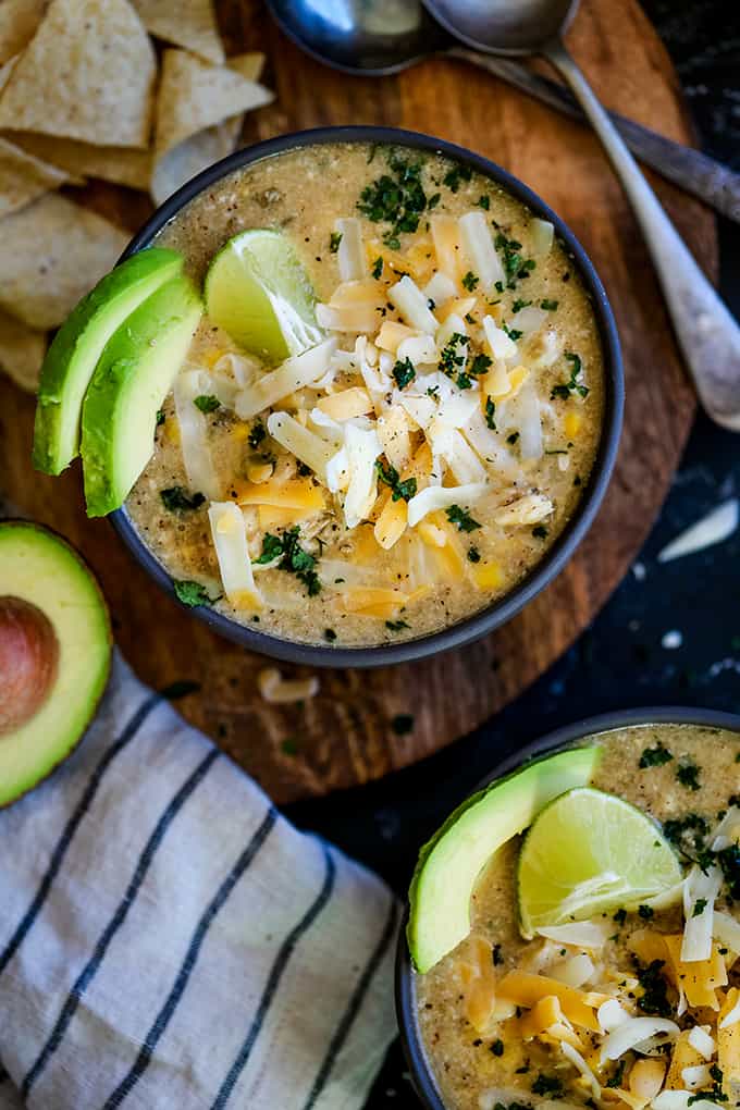 Two bowls of Slow Cooker Coconut Lime White Chicken Chili sit on a wooden surface surrounded by tortilla chips, a striped napkin, avocado half and two spoons.