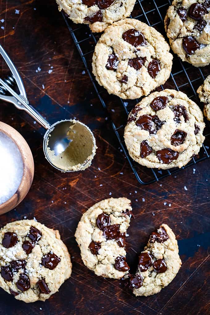 Salted Chocolate Chunk Cookies sit on a cooling rack and on a dark surface surrounded by a bowl of sea salt and a cookie scoop.