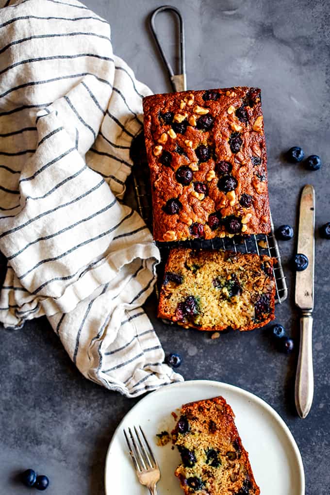 A loaf of Blueberry Walnut Banana Bread flanked by a striped napkin, knife and a plate with a piece of bread on it.